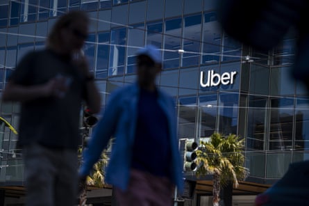 People walk in shadow in front of a tall glass building which bears the Uber logo on the front in white lettering.