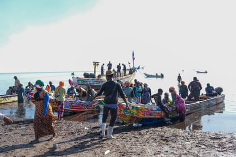 Colourful boats full of people line up in the shallows of the sea. Other people stand next to them in the water and on the beach. Other boats can be seen out in the water