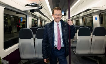 Andy Street wearing pink tie standing inside an empty train holding on to a pole in an aisle.