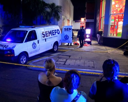 A night shot of three onlookers standing behind a police cordon