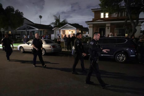 Uniformed law enforcement officers walk down a street at dusk while people look on from their lighted homes.