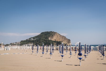 A sandy beach with rows of closed blue and white parasols and a headland in the background