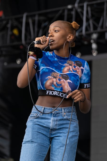 A woman of colour holding a microphone and singing, wearing a blue top with angels on it and faded blue jeans
