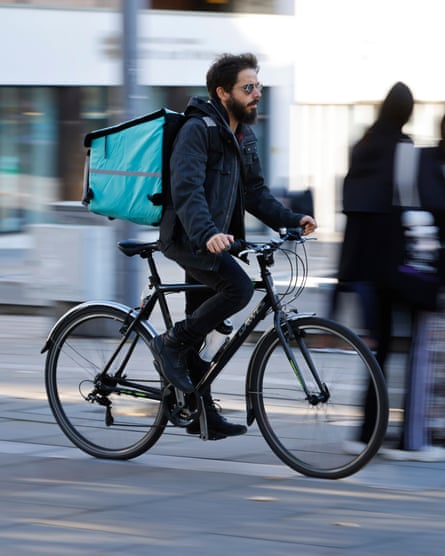 Man wearing dark clothes with an insulated food bag on his back cycles on paving stones