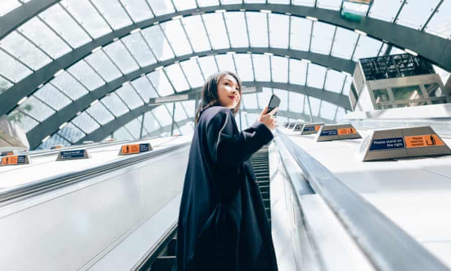 Low angle view of confident Asian woman checking emails on smart phone, riding an escalator, commuting to work.