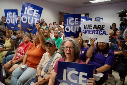 group of people seated inside, many holding blue signs saying 'ICE out' or 'we are America'