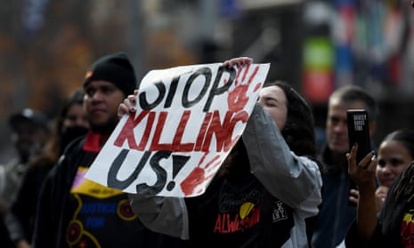 Woman wearing a shirt with the Aboriginal flag holding up a sign that says 'stop killing us!'