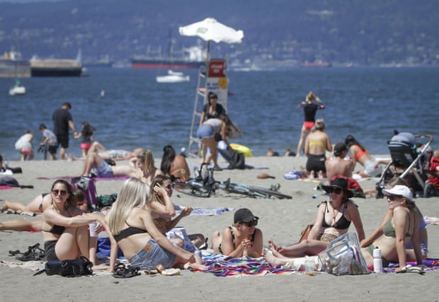 People sit on the sand on a sunny day at the beach. A group of women in the foreground sit on towels in the sand, propping themselves up on their arms.