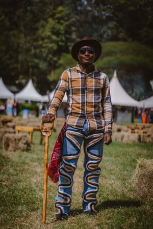 A man wears a plaid shirt, cowboy hat, sunglasses and blue jeans with serpent detailing holds a cane