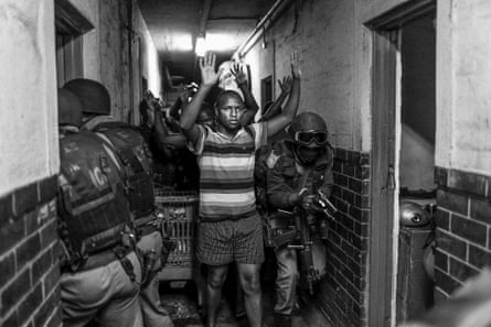 Monochrome picture of a young man in a hallway with his hands above his head as police enter rooms around him