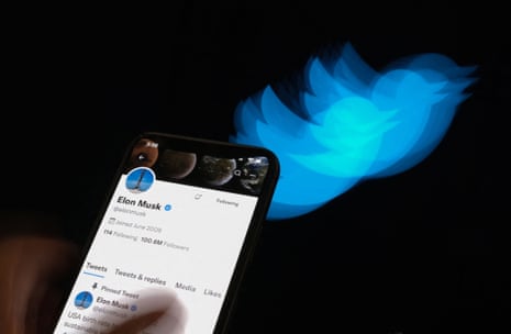Photo illustration of a blurred blue Twitter logo that appears to be flying off to the right. In the foreground, a hand holds a smartphone with Elon Musk's Twitter profile showing.