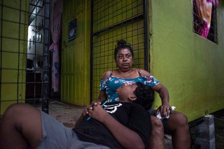 Um menino mesoamericano e uma mulher de meia-idade, com ele encostado nas pernas dela e olhando para ela enquanto ela olha para longe