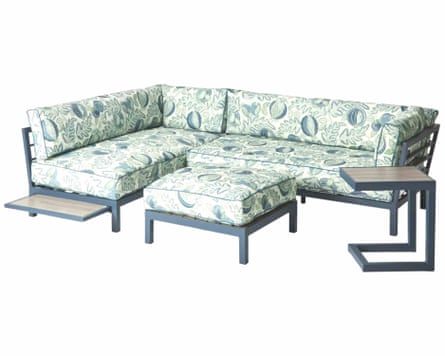 An L-shapes sofa with a dark grey fame. The Cushions have a botanical green-blue and white pattern. There is a large footstall in front of the sofa with a dark grey frame and a cushion that has a botanical pattern in white green and dark blue. There is a side table to right edge of the sofa. This has a dark grey angular c-shaped frame and a wooden top.