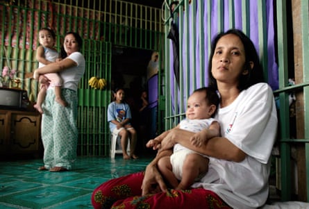 A woman sits next to a wall in a cell with bars with a baby on her lap, while another woman stands in the room behind her holding a baby. A third woman sits on a plastic stool by the door.
