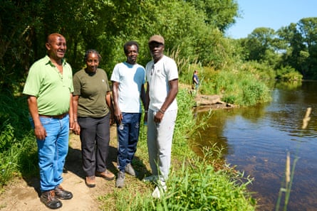 Three men and a woman stand close together on a leafy riverside path on a sunny day