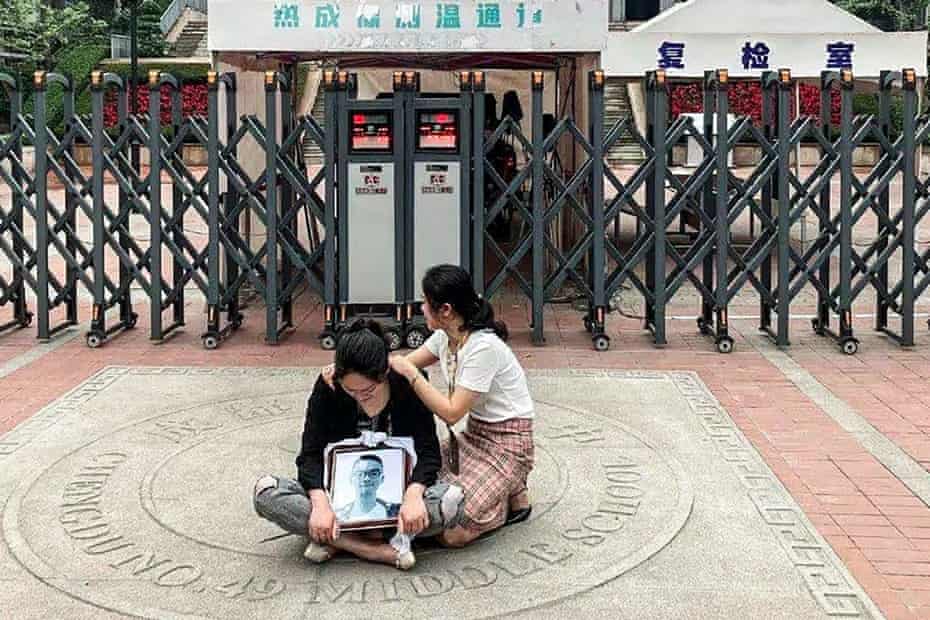 A woman sitting cross-legged on the ground in tears in front some gates, holding a framed picture of a teenager, with another woman consoling her. Chinese characters of the school can be seen displayed above the entrance