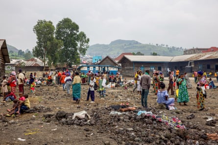 An open area full of people with latrines and classrooms in the background