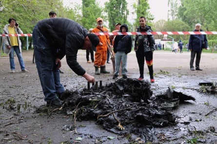 A man leans over charred, black fragments of a drone lying in a muddy puddle as others look on from behind red and white tape.