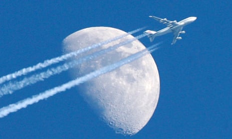 A jumbo jet flies in front of the moon, leaving a long trail of vapour