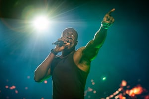 Stormzy in a black vest stands pointing and rapping into a microphone under a bright light