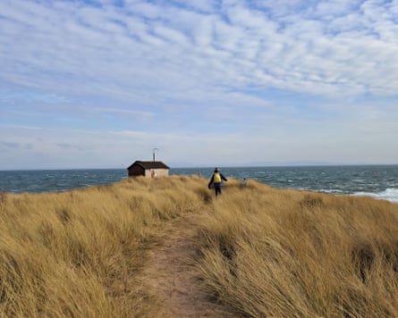flat sand dune going into a distant point – featuring a lone person and a hut – with the sea and a mackerel sky behind it
