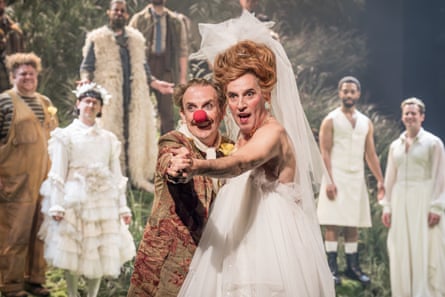 Two men, one wearing a red nose, the other dressed as a woman in a wedding dress, dance together in hold