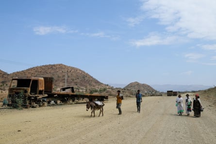 People with donkeys walk along a dusty road past burnt-out trucks