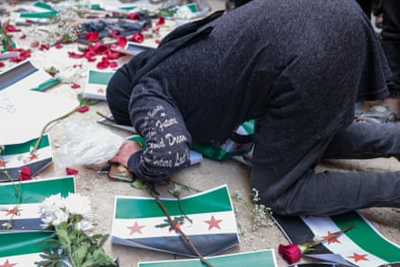 A woman prays on the ground which is covered with roses and Syrian flags