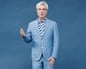 David Byrne in a blue suit and striped shirt, with his hand outstretched