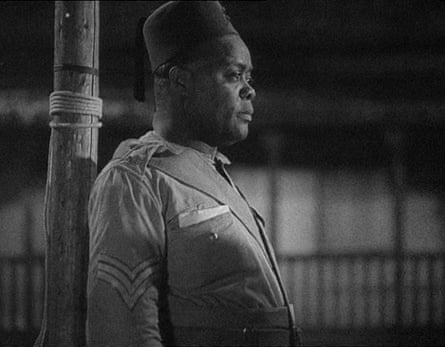 A medium closeup of a man in profile standing in repose against a pole or mast