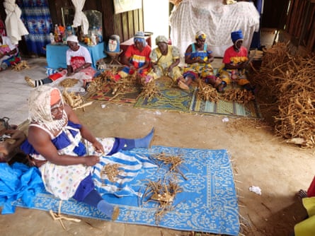 A woman sitting on a rug works on a root with a knife. Five other women doing similar work sit in the same room