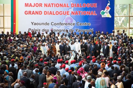 The group of officials stands in front of a large crowd of people. Behind them a banner reads: ‘Major national dialogue. Yaounde conference centre.