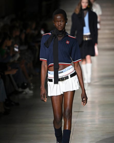 A model wears a white pleated mini skirt, with the waistband of shiny light blue pants visible above. She is also wearing a polo-style crop top and black scarf