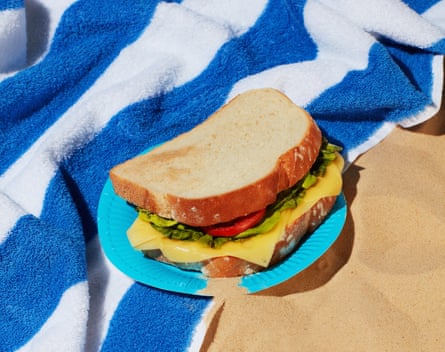 Paper plate with sand and a sandwich on it, on blue and white striped towel on the beach