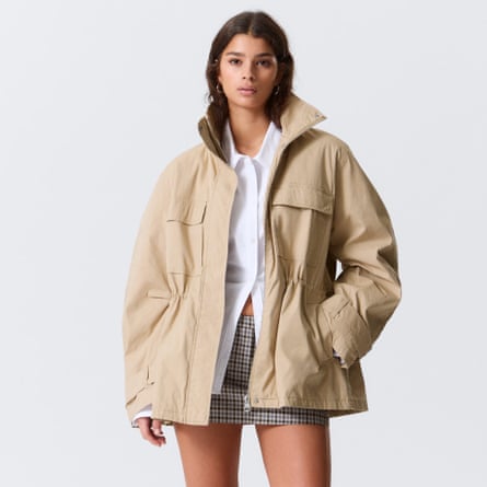 A model wears the Weekday cinched waist canvas jacket in oateal. They wear it over a white shirt and checked miniskirt