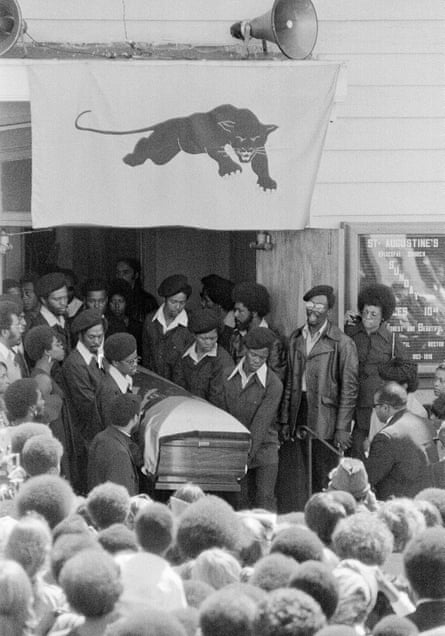 Black and white image of men in matching berets and butterfly collars carrying a casket into a crowd.