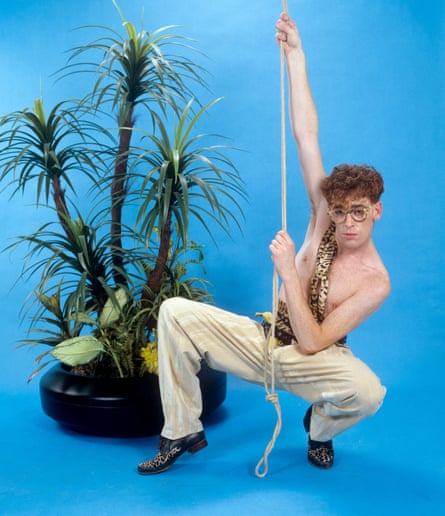 James crouches and holds a rope as if swinging, wearing chinos with a leopardskin scarf over one shoulder and across his bare chest, in front of a potted plant