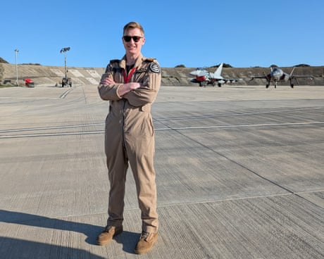 Pilot stands with arms folded on airfield with fighter jets in the background