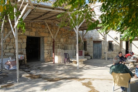 two men sit in the sunshine at a table in front of stone buildings with a wooden canopy, overhanging trees and stacks of empty crates in front. A red motorbike is parked in the background.