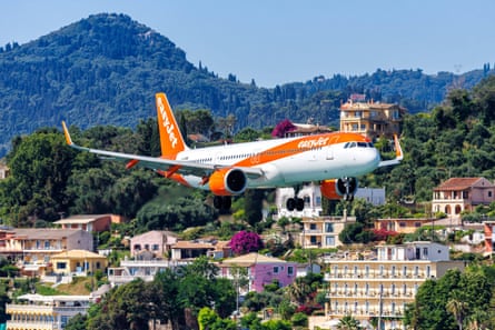 An easyJet Airbus 321 flies near Corfu airport with a scenic view of houses and mountains on the island behind it.