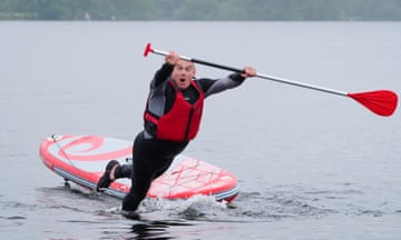 Ed Davey mid-fall from a paddleboard into a lake. He is falling forwards, holding the paddle in front of him and looking at the camera. He is wearing a red buoyancy aid
