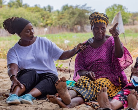 Three black women sit on the ground. A younger woman holds a microphone in front of an older woman who is pointing into the distance as another older woman listens while holding another mic