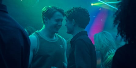 Two young men face each other closely in a club setting with teal and purple lighting