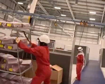 People dressed in orange with white helmets inside a factory, one seen pulling down metallic shelves.