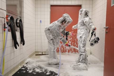 A nurse, already covered in white foam, sprays the foam at another nurse. Both are wearing protective clothing and are in a white-tiled room
