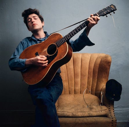 Bob Dylan sitting on the arm of a chair, playing his guitar, New York City, 1964