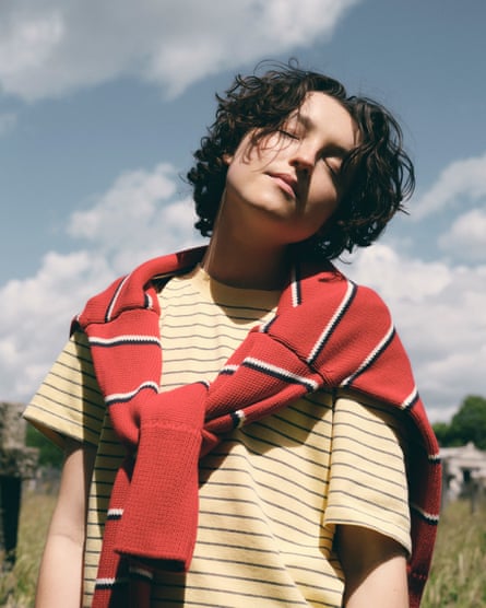 Bella Ramsey wearing a stripy yellow T-shirt with a stripy red jumper over her shoulders, standing with her eyes clothes and her head slightly tipped to her left
