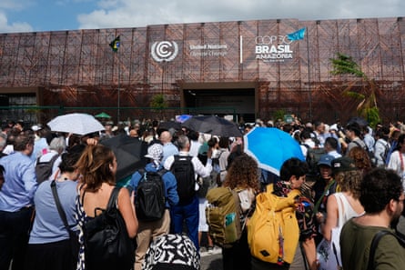 A large crowd of people, some holding parasols to shield themselves from the sun, stand outside the Cop30 venue