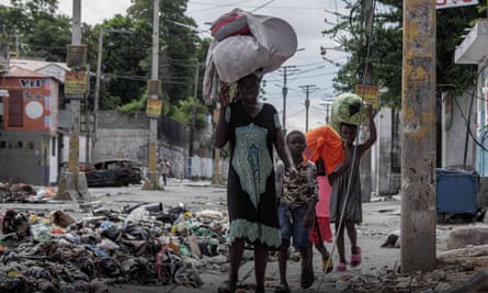 An African-Caribbean woman and three children carry bundles of belongings as they walk past piles of rubbish in a street with a burned-out car int the background
