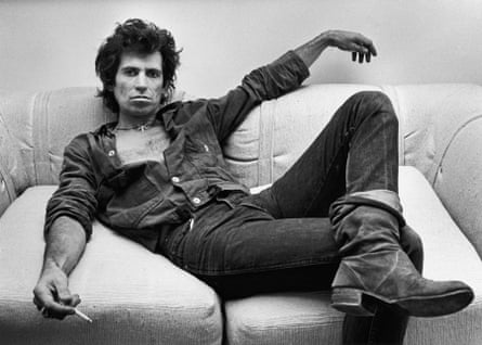 A man dressed in black stretched over a sofa with a cigarette in his right hand